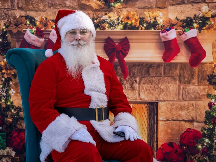 Santa seated in a teal armchair in front of a stone fireplace with stockings and garland