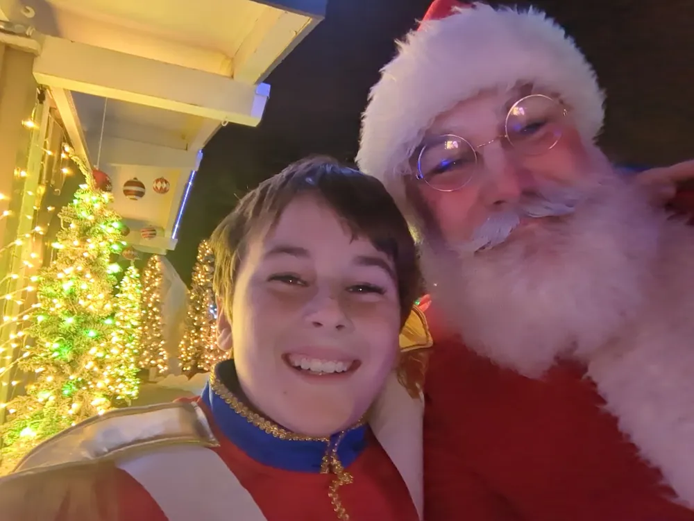 Santa smiling for a selfie with a boy dressed as a toy soldier in front of lit Christmas trees