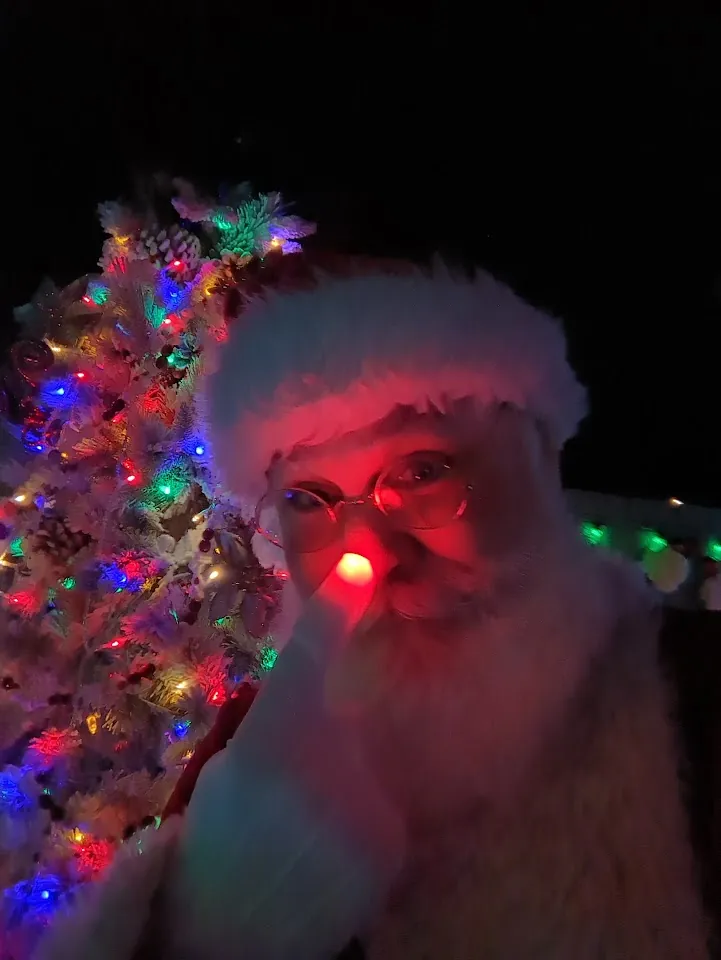 Santa in the dark beside a lit Christmas tree, with a red glow at his nose