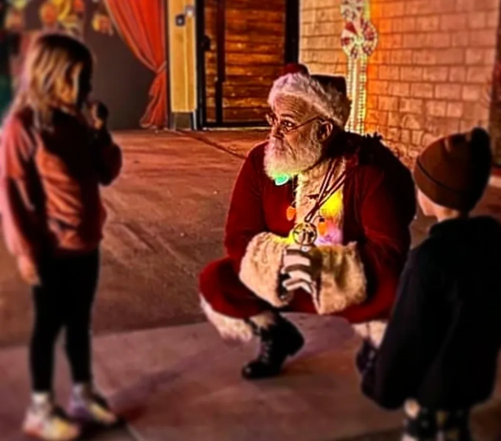 Santa crouching on a sidewalk at night to talk with two small children, wearing a string of colored lights