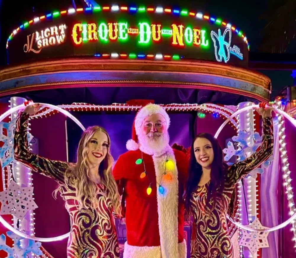 Santa standing between two performers in sparkling gold and red costumes, each holding a lit hoop, under a marquee sign
