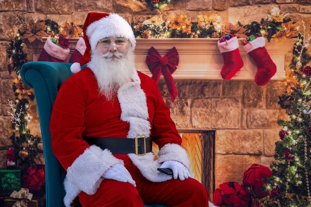 Santa seated in a teal armchair in front of a stone fireplace with stockings and garland